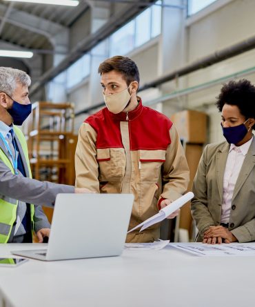 Woodworking factory laborer and quality control inspectors wearing protective face masks while talking and analyzing paperwork at production facility.