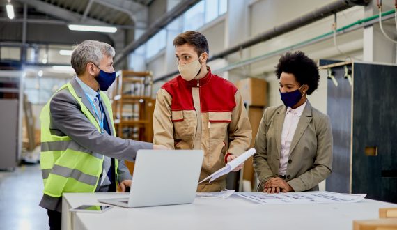 Woodworking factory laborer and quality control inspectors wearing protective face masks while talking and analyzing paperwork at production facility.